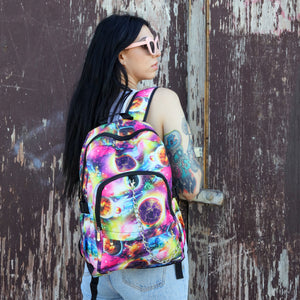 Model is standing in front of a painted brown wall wearing the pink and purple space galaxy vegan backpack. The backpack is facing forward to highlight the multicoloured space/planet/galaxy print, two zip pockets, two elastic side pockets and detachable silver chain.