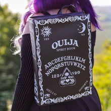 Load image into Gallery viewer, The gothx ouija spirit book vegan backpack modelled by Azariah. They are facing away from the camera with the backpack on their back facing the camera   to highlight the embroidered planchette and white printed detailing featuring a ouija board, pentagrams and lace pattern.
