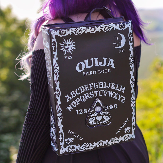The gothx ouija spirit book vegan backpack modelled by Azariah. They are facing away from the camera with the backpack on their back facing the camera   to highlight the embroidered planchette and white printed detailing featuring a ouija board, pentagrams and lace pattern.