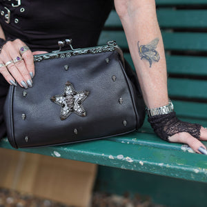 The GothX skull struck vegan vintage clasp handbag being held by a model on a bench. The bag is facing forward to highlight the vintage ball clasp close, floral metal top detailing, detachable handle strap, skull studs and skull star with chains, studs and crystals.