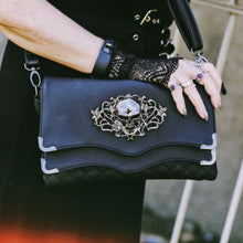 Load image into Gallery viewer, The GothX Skulls and Roses Quilted Clutch Bag being held by an alternative witchy styled goth model wearing it on her shoulder. The vegan leather clutch bag is facing forward and being opened to highlight the two magnetic clip close flaps with metal corners, a stitch quilted front, a skulls and roses metal centrepiece and two D rings on either side for a detachable strap. The mini bag is inspired by gothic grunge witchy fashion.
