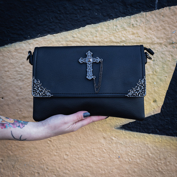 The GothX Don't cross me vegan oversized clutch bag being held in front of a graffiti wall. Bag is facing forward to highlight the metal lace effect detailing on the front flap corners and the studded cross with chain applique.