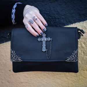 The GothX Don't cross me vegan oversized clutch bag being held in front of a graffiti wall. Bag is facing forward to highlight the metal lace effect detailing on the front flap corners and the studded cross with chain applique.