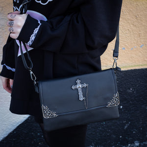 The GothX don't cross me vegan oversized clutch bag hanging on a model's shoulder with the detachable adjustable shoulder strap. Bag is facing forward to highlight the metal lace effect detailing on the front flap corners and the studded cross with chain applique.