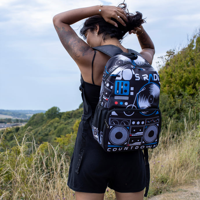 Meera is stood in a grass field wearing the retro black 80s stereo vegan backpack. The bag is facing the camera to highlight the 80s stereo boombox front print, front zip pocket, main zip compartment, two side pockets and top handle.