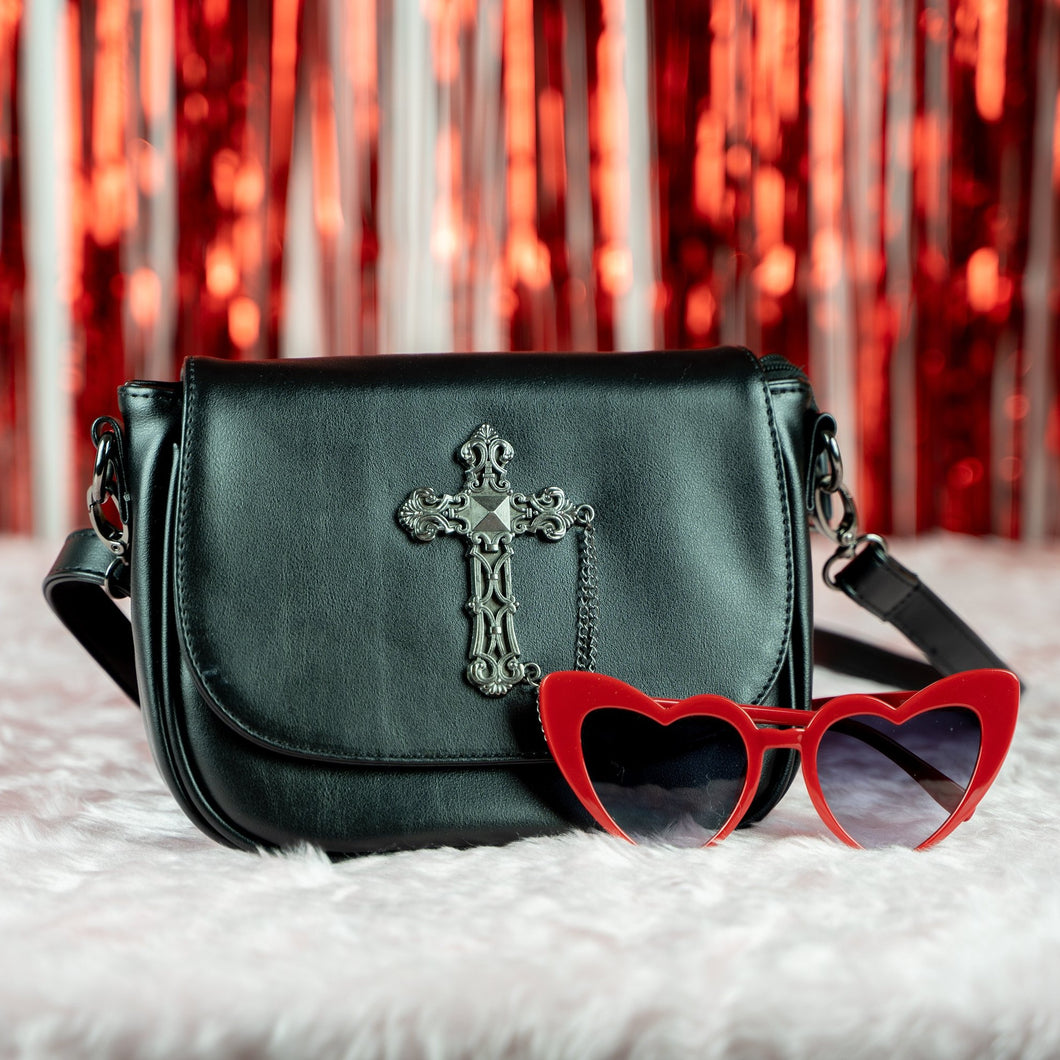 The gothx don't cross me vegan shoulder bag on white faux fur, with a red tassel background behind and next to red heart sunglasses. The bag is facing forward to highlight the studded cross chain centrepiece.
