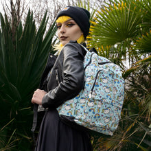 Load image into Gallery viewer, Claudia with mid length yellow hair is stood in front of a green plant area wearing an all black outfit and black beanie with the kawaii unicorn vegan backpack on one shoulder. She is facing away from the camera looking over her shoulder and smiling. The bag is facing towards the camera to highlight the pastel blue rainbow unicorn print, two front zip pockets and detachable silver chain.
