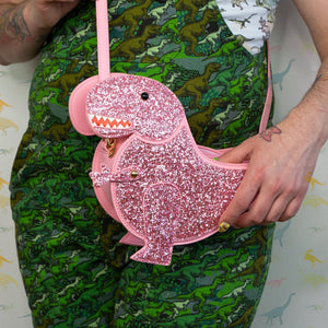 Model wearing green dinosaur dungarees posing with the kawaii pink glitter dino vegan bag. The model is wearing run and fly dinosaur tshirt with green dinosaur dungarees whilst the pink dinosaur bag rests on their shoulder. The bag is facing forward to highlight the pastel pink glittered side, dinosaur face, gold metal detailing, detachable strap and moveable arm.