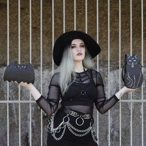 Model with silver hair wearing a goth outfit is holding the gothx pagan black cat vegan shoulder bag out on their right hand and the gothx bat bag on their left hand. The bag is facing forward to highlight the embroidery details including a pentagram collar, yellow eyes and paws.