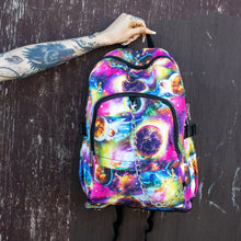 Load image into Gallery viewer, Model is holding the pink and purple space galaxy vegan backpack in front of a painted brown wall. The backpack is facing forward to highlight the multicoloured space/planet/galaxy print, two zip pockets, two elastic side pockets and detachable silver chain.
