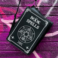 Load image into Gallery viewer, The GothX Book of Spells Vegan Messenger Bag. Vegan black leather with white witch pagan magic symbols printed on the front and spine of the 3d book bag. Bag is being held up in front of a pink and purple graffiti wall.
