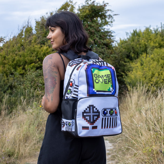 Meera is stood in a grass field wearing an all black outfit with the white game over vegan backpack on her back. The backpack features a 90s inspired gameboy print with buttons and a screen saying game over.
