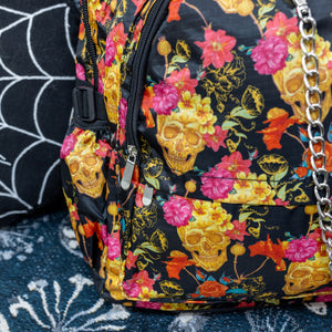 Close up of the floral gold skull nylon chain vegan backpack sat on a spooky halloween blanket next to a spider web cushion. The backpack is facing forward to highlight the gold skulls, pink and red flowers print on a black background, the two front zip pockets and two side pockets with a silver decorative chain.