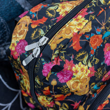 Load image into Gallery viewer, Close up of the floral gold skull nylon chain vegan backpack sat on a spooky halloween blanket next to a spider web cushion. The backpack is facing forward to highlight the gold skulls, pink and red flowers print on a black background, the two front zip pockets and two side pockets with a silver decorative chain.
