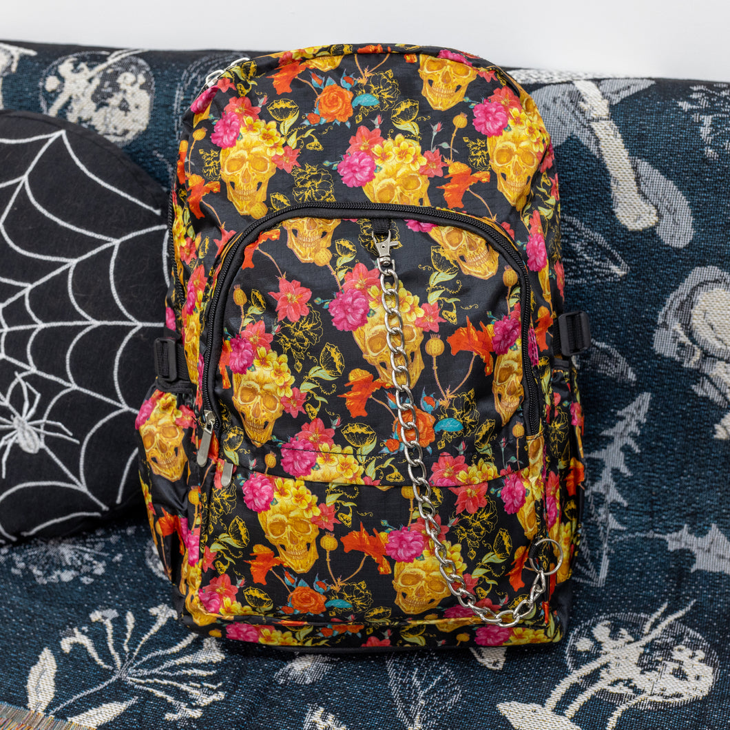 The floral gold skull nylon chain vegan backpack sat on a spooky halloween blanket next to a spider web cushion. The backpack is facing forward to highlight the gold skulls, pink and red flowers print on a black background, the two front zip pockets and two side pockets with a silver decorative chain.
