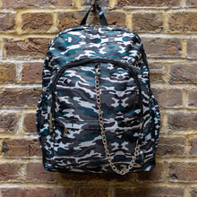 Load image into Gallery viewer, Forest Camouflage vegan backpack with chain hanging on a brick wall. The backpack with a dark green and brown camo print is facing forward highlighting the two front zip pockets, two side pockets and silver decorative chain.

