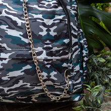 Load image into Gallery viewer, Close up of the Forest Camouflage vegan backpack with chain sat outside in front of a tropical plant and brick wall. The backpack with a dark green, brown camo print is facing forward highlighting the two front zip pockets, two side pockets and silver decorative chain.
