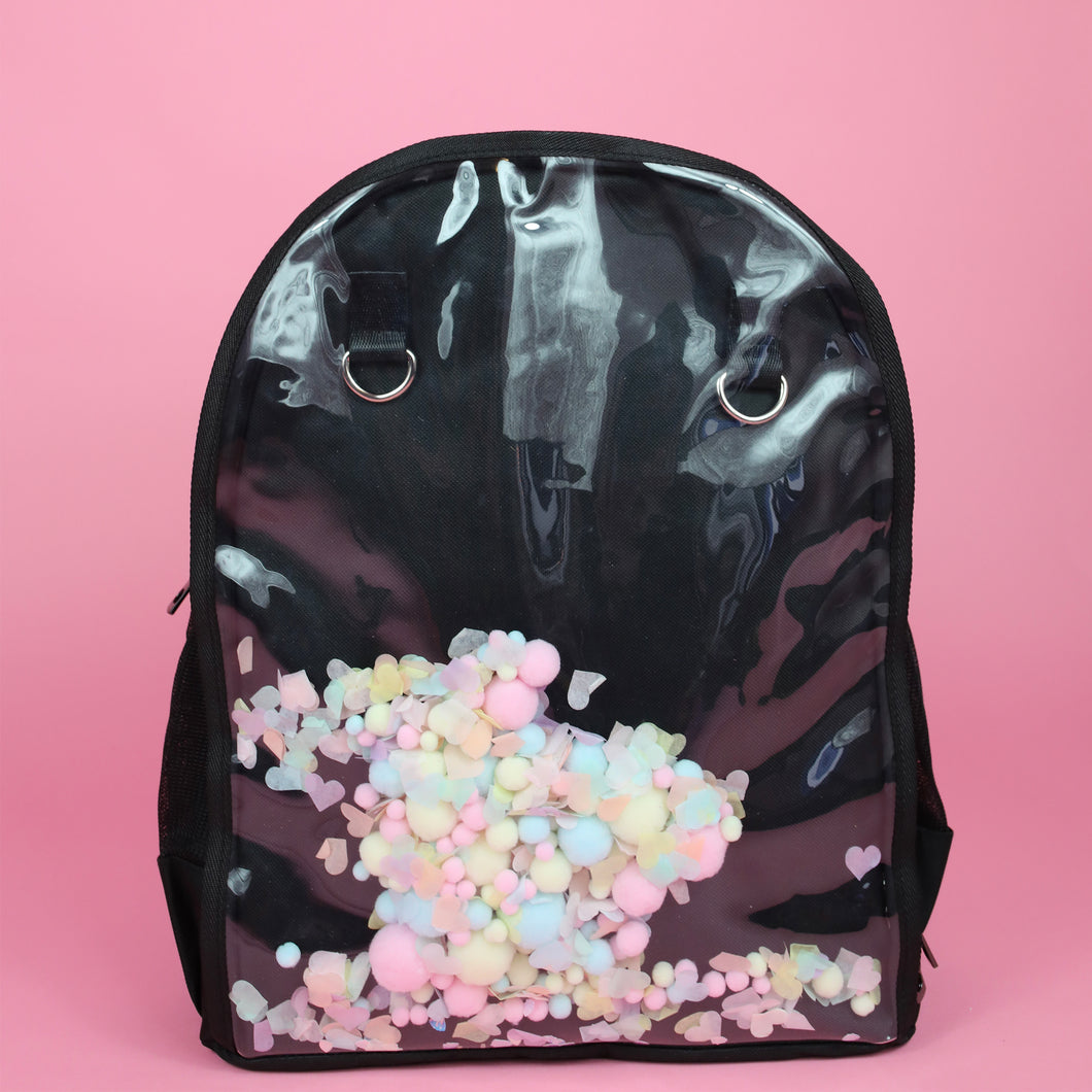 The Kawaii Black & Clear Window Vegan Ita Backpack on a pastel pink background. The bag is facing forward filled with pastel confetti and pom poms to highlight the clear display window and inside D rings to hang things from.