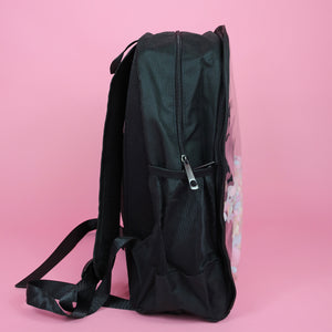 The Kawaii Black & Clear Window Vegan Ita Backpack on a pastel pink background. The bag is facing right filled with pastel confetti and pom poms to highlight the clear display window, double zip opening, side pockets and padded adjustable shoulder straps.
