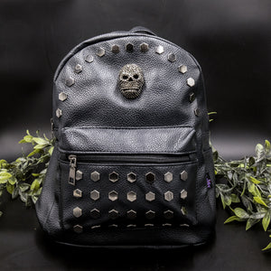 The Gothx skull head large studs vegan mini backpack on a black background with foliage surrounding it. The bag is facing forward to highlight the large hexagonal flat studded front zip pocket, diamante effect silver skull and studs along the top zip line. 