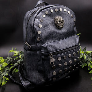 The Gothx skull head large studs vegan mini backpack on a black background with foliage surrounding it. The bag is facing forward angled right to highlight the large hexagonal flat studded front zip pocket, diamante effect silver skull and studs along the top zip line. 
