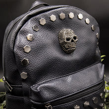 Load image into Gallery viewer, Close up of the front of the gothx skull head large studs vegan mini backpack showing the diamante effect skull and large hexagonal flat studs along the top zip line. The bag is facing forward angled to the right on a black background with foliage.
