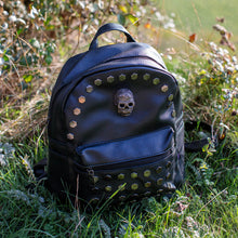 Load image into Gallery viewer, The GothX skull head large studs vegan mini backpack sat on a grass field. The bag is facing forward to highlight the large hexagonal flat studded front zip pocket, diamante effect silver skull and studs along the top zip line.
