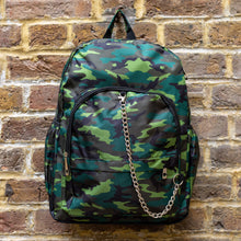 Load image into Gallery viewer, The Jungle Camouflage Backpack hanging outside on a brick wall. The green, brown, khaki and black vegan friendly backpack is facing forward to highlight the two front zip pockets with a silver draping detachable chain, the two side elasticated pockets, the top handle and the main double zip compartment.
