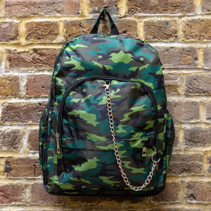 The Jungle Camouflage Backpack hanging outside on a brick wall. The green, brown, khaki and black vegan friendly backpack is facing forward to highlight the two front zip pockets with a silver draping detachable chain, the two side elasticated pockets, the top handle and the main double zip compartment.