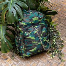 Load image into Gallery viewer, The Jungle Camouflage Backpack sat outside on a brick floor surrounded by giant monstera leaves and other green foliage. The green, brown, khaki and black vegan friendly backpack is sat facing forward to highlight the two front zip pockets with a silver draping detachable chain, the two side elasticated pockets, the top handle and the main double zip compartment.
