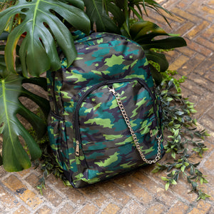 The Jungle Camouflage Backpack sat outside on a brick floor surrounded by giant monstera leaves and other green foliage. The green, brown, khaki and black vegan friendly backpack is sat facing forward to highlight the two front zip pockets with a silver draping detachable chain, the two side elasticated pockets, the top handle and the main double zip compartment.
