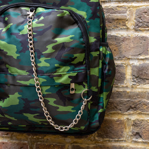 Close up of the Jungle Camouflage Backpack hanging outside on a brick wall. The green, brown, khaki and black vegan friendly backpack is facing forward to highlight the two front zip pockets with a silver draping detachable chain, the two side elasticated pockets, the top handle and the main double zip compartment.