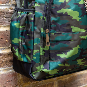 Close up of the Jungle Camouflage Backpack hanging outside on a brick wall. The green, brown, khaki and black vegan friendly backpack is facing forward to highlight the two front zip pockets with a silver draping detachable chain, the two side elasticated pockets, the top handle and the main double zip compartment.