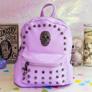 The GothX Pastel Lilac Skull Head Small Studs Vegan Mini Backpack sat on an iridescent pink background with a palmistry guide poster, phrenology candle, two skulls and purple pumpkins in the background. The bag is facing forward to highlight the dark grey metal stud detailing, diamanté effect skull, front zip pocket, two side slip pockets and top handle.