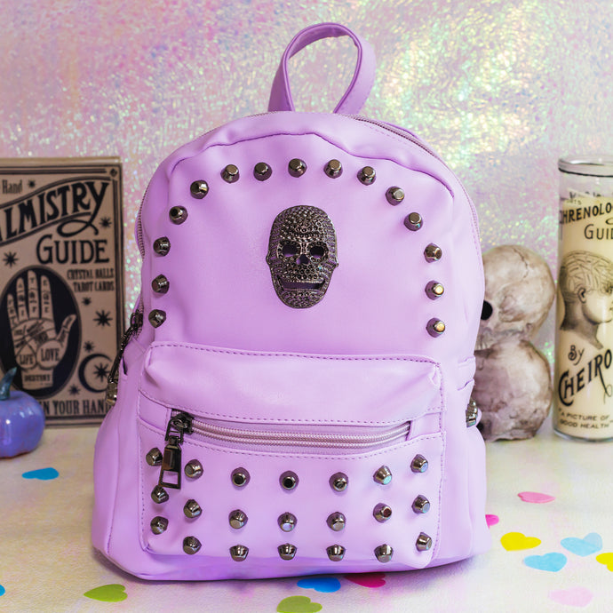 The GothX LIMITED EDITION Pastel Lilac Skull Head Small Studs Vegan Mini Backpack sat on an iridescent pink background with a palmistry guide poster, phrenology candle, two skulls and purple pumpkins in the background. The bag is facing forward to highlight the dark grey metal stud detailing, diamanté effect skull, front zip pocket, two side slip pockets and top handle.