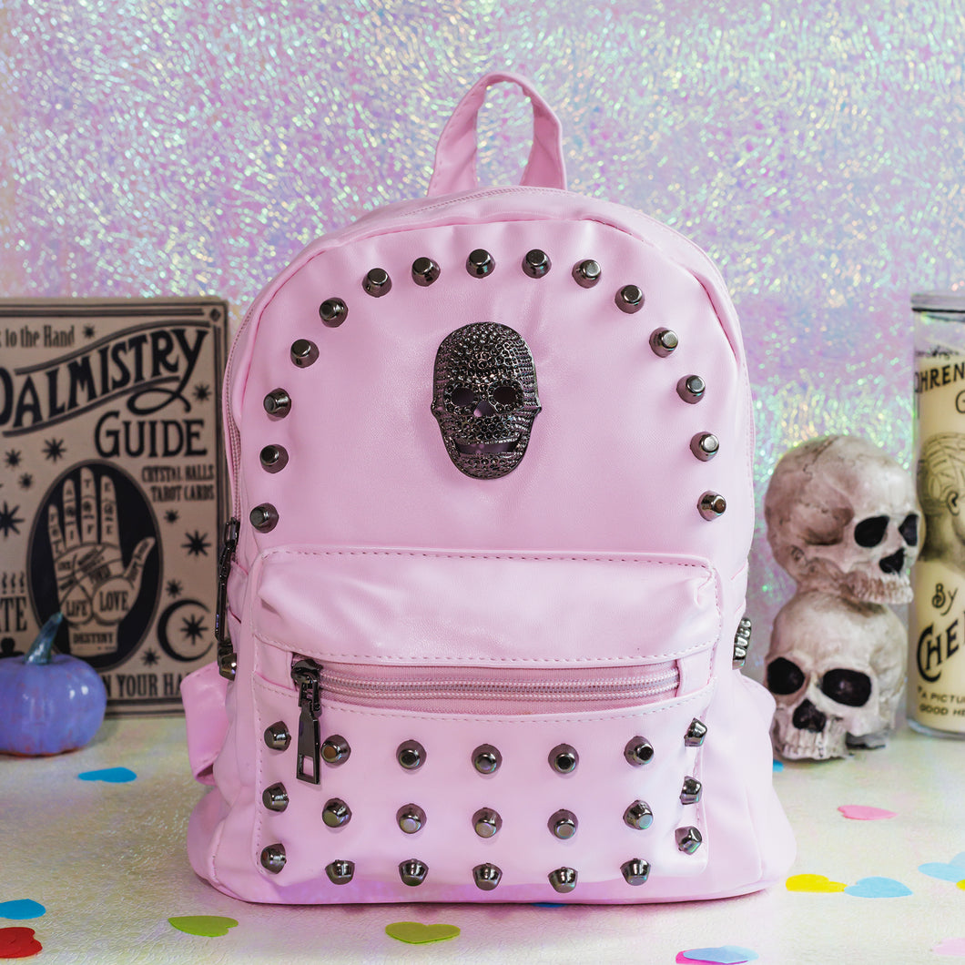 The GothX Pastel Pink Skull Head Small Studs Vegan Mini Backpack sat on an iridescent pink background with a palmistry guide poster, phrenology candle, two skulls and purple pumpkins in the background. The vegan leather bag is facing forward to highlight the dark grey metal stud detailing, diamanté effect skull, front zip pocket, two side slip pockets and top handle.