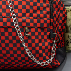 Close up of the Red Checkerboard Backpack sat on a grey background with a palmistry guide book and two skull stack on the right and a phrenology guide candle and three skull stack on the left. The vegan friendly bag is facing forward to highlight the red and black check print, two front zip pockets, two elasticated side pockets, main top double zip pocket and silver draping decorative chain.