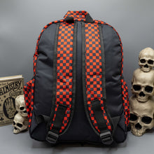 Load image into Gallery viewer, The Red Checkerboard Backpack sat on a grey background with a palmistry guide book and two skull stack on the right and a phrenology guide candle and three skull stack on the left. The vegan friendly bag is facing away from the camera to highlight the plain black back, the two side elasticated pockets, the top handle and the two adjustable padded shoulder straps.
