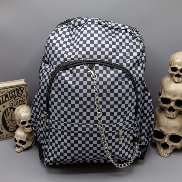  The Grey Checkerboard Backpack sat on a grey background with a palmistry guide book and two skull stack on the right and a phrenology guide candle and three skull stack on the left. The vegan friendly bag is facing forward to highlight the grey and black check print, two front zip pockets, two elasticated side pockets, main top double zip pocket and silver draping decorative chain.