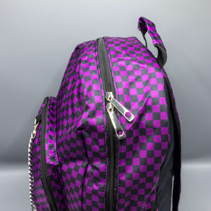 Close up of the Purple Checkerboard Backpack sat on a grey background. The vegan friendly bag is facing forward to highlight the purple and black check print, two front zip pockets, two elasticated side pockets, main top double zip pocket and silver draping decorative chain.