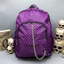 Load image into Gallery viewer, The Purple Checkerboard Backpack  sat on a grey background with a palmistry guide book and two skull stack on the right and a phrenology guide candle and three skull stack on the left. The vegan friendly bag is facing forward to highlight the purple and black check print, two front zip pockets, two elasticated side pockets, main top double zip pocket and silver draping decorative chain.
