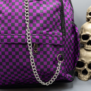 Close up of the Purple Checkerboard Backpack sat on a grey background with a palmistry guide book and two skull stack on the right and a phrenology guide candle and three skull stack on the left. The vegan friendly bag is facing forward to highlight the purple and black check print, two front zip pockets, two elasticated side pockets, main top double zip pocket and silver draping decorative chain.
