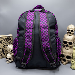 The Purple Checkerboard Backpack sat on a grey background with a palmistry guide book and two skull stack on the right and a phrenology guide candle and three skull stack on the left. The vegan friendly bag is facing away from the camera to highlight the plain black back, the two side elasticated pockets, the top handle and the two adjustable padded shoulder straps.