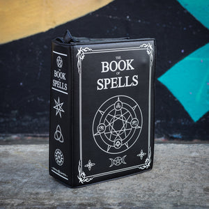 The GothX Book of Spells Vegan Messenger Bag. A book shaped black vegan bag with the book of spells witch pagan white print. Shown angled to the right to show off the pentagram and magic symbol printed spine in front of a graffiti wall.