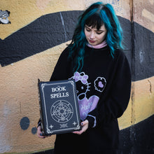 Load image into Gallery viewer, A blue haired model stood in front of a graffiti wall wearing an oversized black jumper whilst holding the GothX book of spells vegan messenger bag down by their side. Vegan black leather with white witch pagan magic symbols printed on the front and spine of the 3d book bag.
