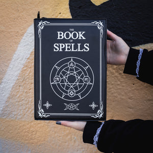 The GothX Book of Spells Vegan Messenger Bag. Vegan black leather with white witch pagan magic symbols printed on the front and spine of the 3d book bag. Bag held up in front of a graffiti wall to show off the front book of spells pentagram print.