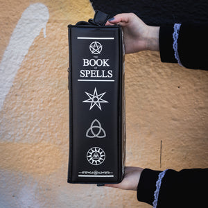 The GothX Book of Spells Vegan Messenger Bag. Vegan black leather with white witch pagan magic symbols printed on the front and spine of the 3d book bag. Bag held up in front of a graffiti wall to show off the side book of spells magical symbol spine print.