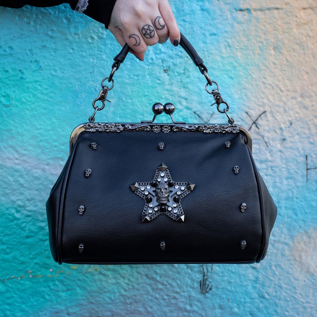 The GothX skull struck vegan vintage clasp handbag being held up in front of a graffiti wall. The bag is facing forward to highlight the vintage ball clasp close, floral metal top detailing, detachable handle strap, skull studs and skull star with chains, studs and crystals.