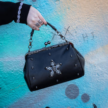 Load image into Gallery viewer, The GothX skull struck vegan vintage clasp handbag being held up in front of a graffiti wall. The vegan leather vintage goth inspired bag is facing forward to highlight the vintage ball clasp close, floral metal top detailing, detachable handle strap, skull studs and skull star with chains, studs and crystals.
