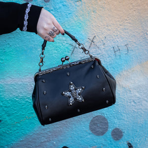 The GothX skull struck vegan vintage clasp handbag being held up in front of a graffiti wall. The vegan leather vintage goth inspired bag is facing forward to highlight the vintage ball clasp close, floral metal top detailing, detachable handle strap, skull studs and skull star with chains, studs and crystals.